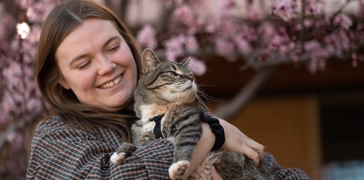 Woman holding tabby cat in front of tree with pink flowers